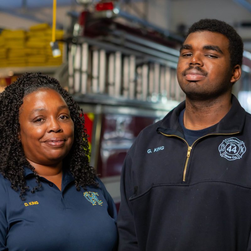 Two firefighters in uniform in front of a fire truck inside the station